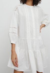 Robe en lin blanche avec un col rond, un devant boutonné et des manches longues ornées de détails en dentelle. Jupe évasée et à volants qui apporte du volume.