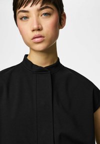 Mlada osoba s kratkom tamnom kosom i plavim očima, odjevena u crnu majicu bez rukava s visokim ovratnikom, na jednobojnoj pozadini.