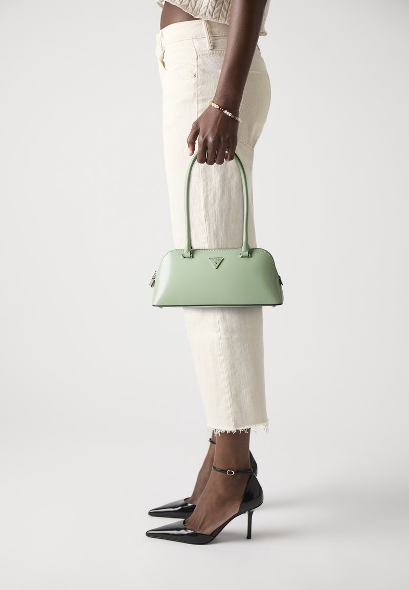 Personne portant un pantalon court blanc et des talons noirs tient à côté d'elle un sac à main structuré vert clair avec deux poignées.