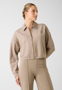 Beżowa, krótka kurtka z zamkiem z przodu, klasycznym kołnierzem i długimi rękawami. Z subtelnym logo na piersi. Gładki, elastyczny materiał.