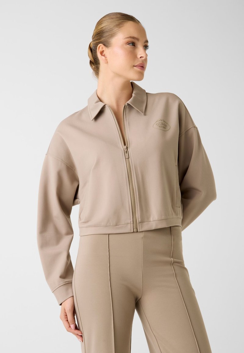 Beżowa, krótka kurtka z zamkiem z przodu, klasycznym kołnierzem i długimi rękawami. Z subtelnym logo na piersi. Gładki, elastyczny materiał.