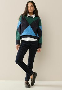 Übergroßer Pullover in grün, blau und schwarz im Argyle-Muster; weißes Hemd mit Kragen darunter; dunkelmarine Cordhosen; glänzende dunkle Schuhe.