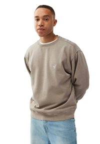 Ung mand med kort hår iført en taupe sweatshirt og lyseblå jeans, stående med hænderne bag ryggen, mod en hvid baggrund.