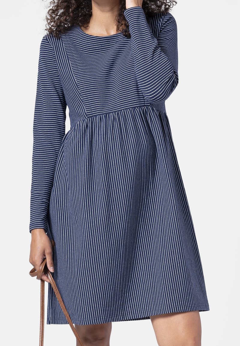 Femme portant une robe bleu marine à manches longues avec des rayures blanches verticales et horizontales, tenant un sac à main à bandoulière en cuir marron sur fond blanc.