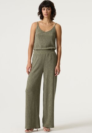 Olivgrüner, strukturierter Jumpsuit mit rund ausgeschnittener Halslinie, dünnen Trägern und lockerem Schnitt. Verjüngtes, weit geschnittenes Bein-Design, getragen mit Riemchensandalen.