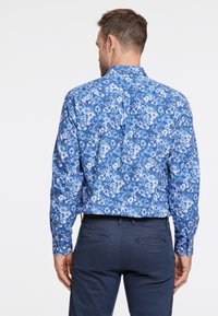 Langärmliges Hemd mit blauen Blumen in verschiedenen Blau- und Weißtönen, mit Knopfkragen und стандартrịȷلتყف. Getragen mit dunklen Hosen.