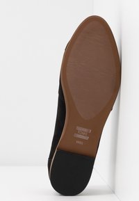 Semelle d'une chaussure TOMS noire avec un dessous marron texturé et un talon noir, marquée W6½, positionnée verticalement contre un mur blanc d'angle.