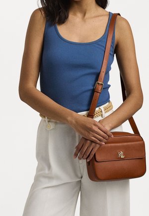 Ruskea nahkainen olkalaukku, jossa on kultainen logo, suorakulmainen muoto, sileä pinta ja säädettävä hihna, joka on henkilön yllä.