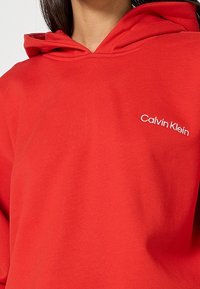 Sweat à capuche rouge avec capuche relevée et logo blanc "Calvin Klein" sur le côté gauche de la poitrine, porté par une personne aux cheveux foncés.