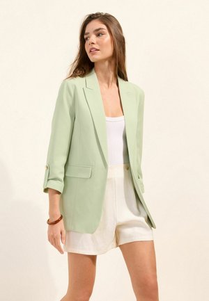 Woman wearing light green blazer, white tank top, and cream shorts, standing against plain light background, looking to her left.