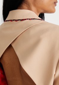 Beiger Stoffkragen mit einem drapierten Design, das mit einer dekorativen Verzierung aus roten und lila Perlen am Saum versehen ist, wodurch die Haut darunter sichtbar wird.