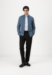 Light blue knitted sweater with a stand collar, zipper closure, and ribbed cuffs, paired with black trousers and black shoes.