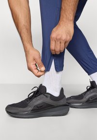 Χέρια που ξεzipάρουν μπλε αθλητικά παντελόνια πάνω από λευκές κάλτσες και μαύρα αθλητικά παπούτσια σε λευκό φόντο.