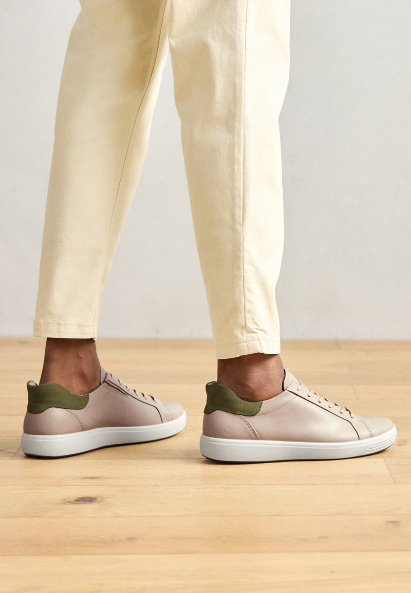 Helle rosa Leder-Sneaker mit einem grünen Fersenakzent, weißer Sohle und sechs Ösen. Getragen mit beigen Hosen, die eine glatte Textur und ein modernes Design zeigen.
