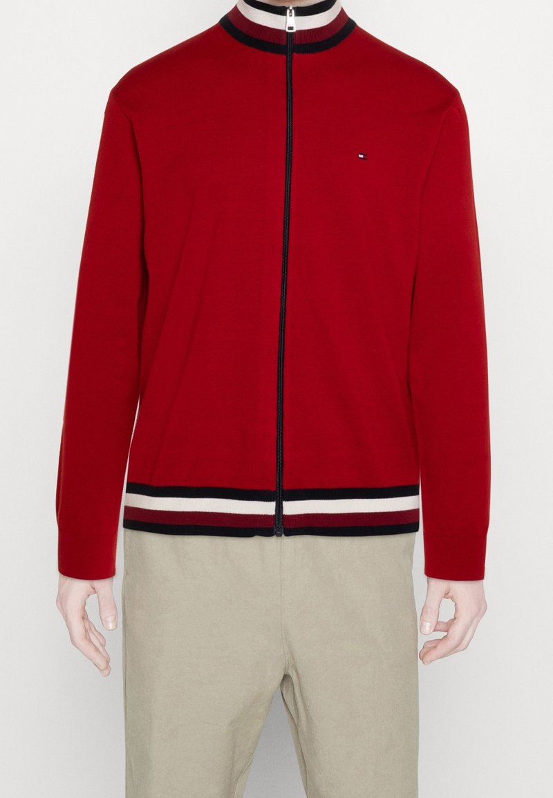 Veste rouge zippée avec col et bas rayés blancs et noirs, portée avec un pantalon beige sur une personne debout sur fond blanc.