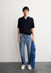 Marineblauw polo met korte mouwen en verticale strepen, gecombineerd met lichtblauwe rechte jeans en witte sneakers, terwijl hij een blauwe jas vasthoudt.