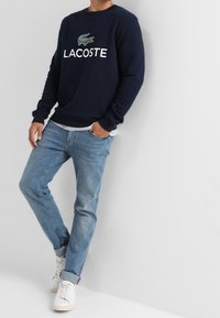Felpa marina con logo "LACOSTE" e design del coccodrillo verde, abbinata a jeans azzurri chiari e scarpe da ginnastica bianche. Tessuto morbido, vestibilità rilassata.
