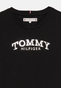 Fekete pamut póló, amely elülső részén ívelt, fényvisszaverő fehér "TOMMY HILFIGER" logóval és csíkos díszítéssel rendelkezik a nyak részen.
