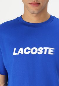 Mėlynas medvilninis marškinėliai su apvaliu kaklu ir ryškiu baltu "LACOSTE" logotipu per krūtinę, parašytu ryškiu, šiek tiek neryškiu šriftu.
