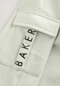 Kremkrāsas audums ar gludu tekstūru, ar taisnstūra kabatu un melnu "BAKER" etiķeti.