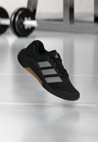 Μαύρο αθλητικό παπούτσι με επάνω μέρος από δίχτυ, τρεις γκρι ρίγες, καουτσούκ σόλα και υφή στο σχέδιο. Διαθέτει μαξιλαρωμένο γιακά και ενισχυμένο μπροστινό μέρος.