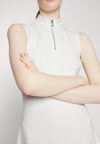 Ärmelloses weißes Kleid mit hohem Kragen und einem silbernen Reißverschluss. Verfügt über ein strukturiertes Muster auf dem Stoff.