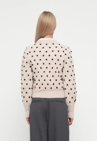 Lys beige strikket genser med svarte prikker, rund hals, og ribbede mansjetter. Kombinert med grå bukser som har en avslappet passform.