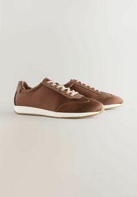 Bruna syntetiska sneakers med beige snörning, slät textur, vit gummisula och en subtil designaccent på hälen.