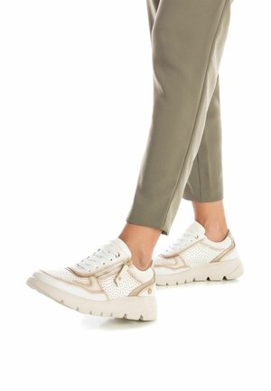 Zapatilla blanca con textura perforada, detalles en color beige y un cierre. Suela de plataforma gruesa. Combinada con pantalones en verde oliva, ajustados.