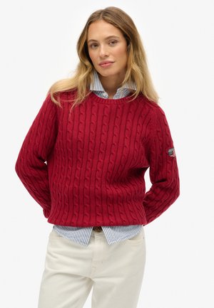 Pull en tricot rouge à col rond, avec des motifs surélevés. Porté par-dessus une chemise rayée bleu clair, associé à un pantalon blanc.