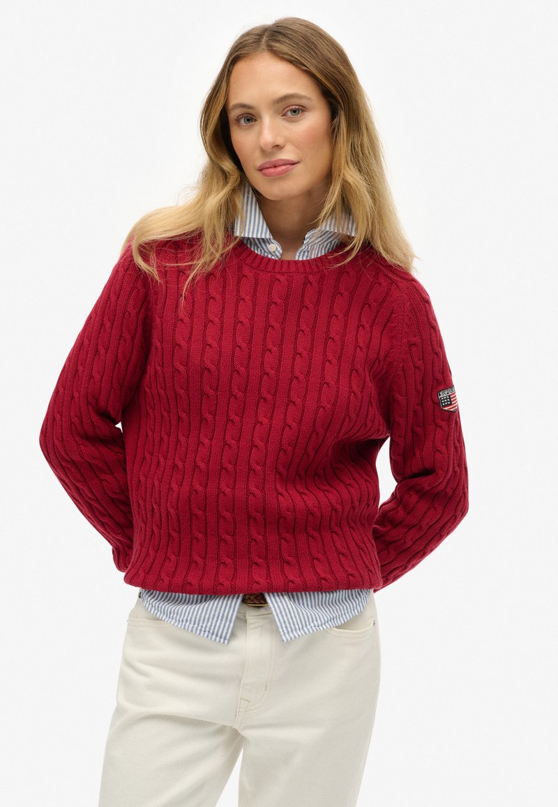 Rode kabelgebreide trui met een ronde hals, voorzien van verhoogde patronen. Draag het over een lichtblauw gestreept shirt, gecombineerd met witte broeken.