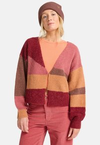 Kleurijke cropped cardigan met geometrische patronen in tinten van rood, roze en bruin. Zachte textuur, geribbelde manchetten en een v-hals ontwerp.