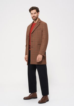 Brązowy w kratę płaszcz z wyłogami, na jeden guzik, z bocznymi kieszeniami, noszony na czerwonym swetrze z dzianiny, zestawiony z czarnymi spodniami i brązowymi botkami.