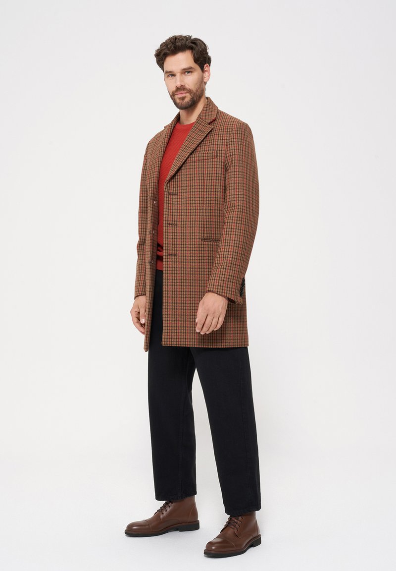 Brązowy w kratę płaszcz z wyłogami, na jeden guzik, z bocznymi kieszeniami, noszony na czerwonym swetrze z dzianiny, zestawiony z czarnymi spodniami i brązowymi botkami.
