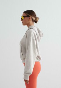 Mujer con cabello castaño recogido en un moño, lleva un sudadera gris claro de gran tamaño, leggings naranjas, pendientes de aro dorados y gafas de sol amarillas reflectantes, mirando hacia la izquierda.