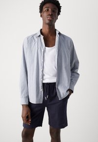 Gestreepte blouse in blauw en wit, gedragen over een witte tanktop; gecombineerd met marineblauwe shorts met een textuurpatroon.