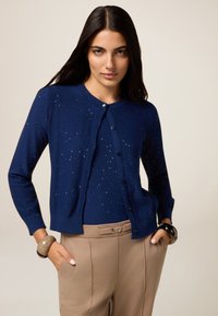 Frau mit langen dunklen Haaren, die eine marineblaue Strickjacke mit Glitzer und beigefarbenen hochgeschnittenen Hosen trägt, Hände in den Taschen, neutraler Hintergrund.
