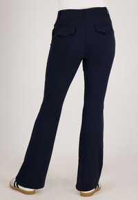 Marinere blauwe stretchbroek met een klokvormig design, voorzien van twee achterzakken en een gladde textuur. G gedragen met witte schoenen en zwarte strepen.