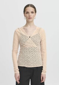 Spitzenoberteil mit langen Ärmeln in sanftem Pfirsich, mit einem Wickeldesign, gewellten Kanten und einem strukturierten Blumenmuster. Kombiniert mit schwarzen Hosen.