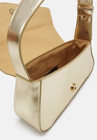 Petit sac à main rectangulaire doré avec rabat et fermeture magnétique, montrant une poche intérieure zippée et une sangle large unique.