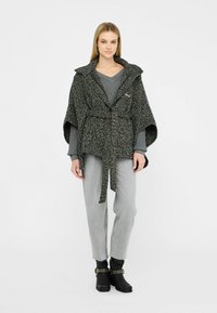 Cappotto stile mantella grigio e nero con una cintura, indossato sopra un maglione grigio e jeans grigio chiaro, abbinato a stivali neri alla caviglia.