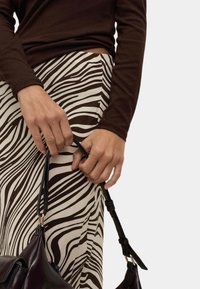 Hnědý top s dlouhým rukávem v kombinaci se smetanovou sukní se vzorem hnědé zebry. Drží tmavě hnědou kabelku s dlouhým popruhem.