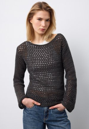 Donna con capelli biondi che indossa un maglione nero a maglia aperta sopra una maglietta bianca e jeans blu, in piedi con le mani nelle tasche.