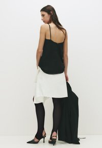 Sort silk camisole med tynde stropper, hvid asymmetrisk nederdel, sorte strømpebukser og spidse sko; enkelt, minimalistisk design med knaplukning på ryggen.
