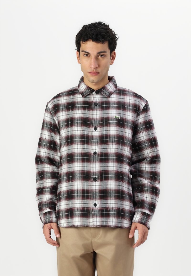 Camisa de cuadros en blanco y negro con acentos en burdeos, con botones en la parte delantera y cuello. Incluye un pequeño logo verde en el pecho. Tejido ligero.