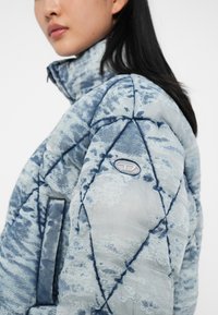 Vrouw met lang zwart haar die een blauw-grijze gewatteerde jas draagt met abstracte patronen en een klein geborduurd logo op de mouw.