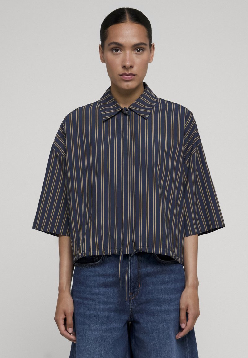 Femme portant une chemise bleu marine à manches larges avec col, à rayures verticales beiges et un jean bleu taille haute sur un fond uni.