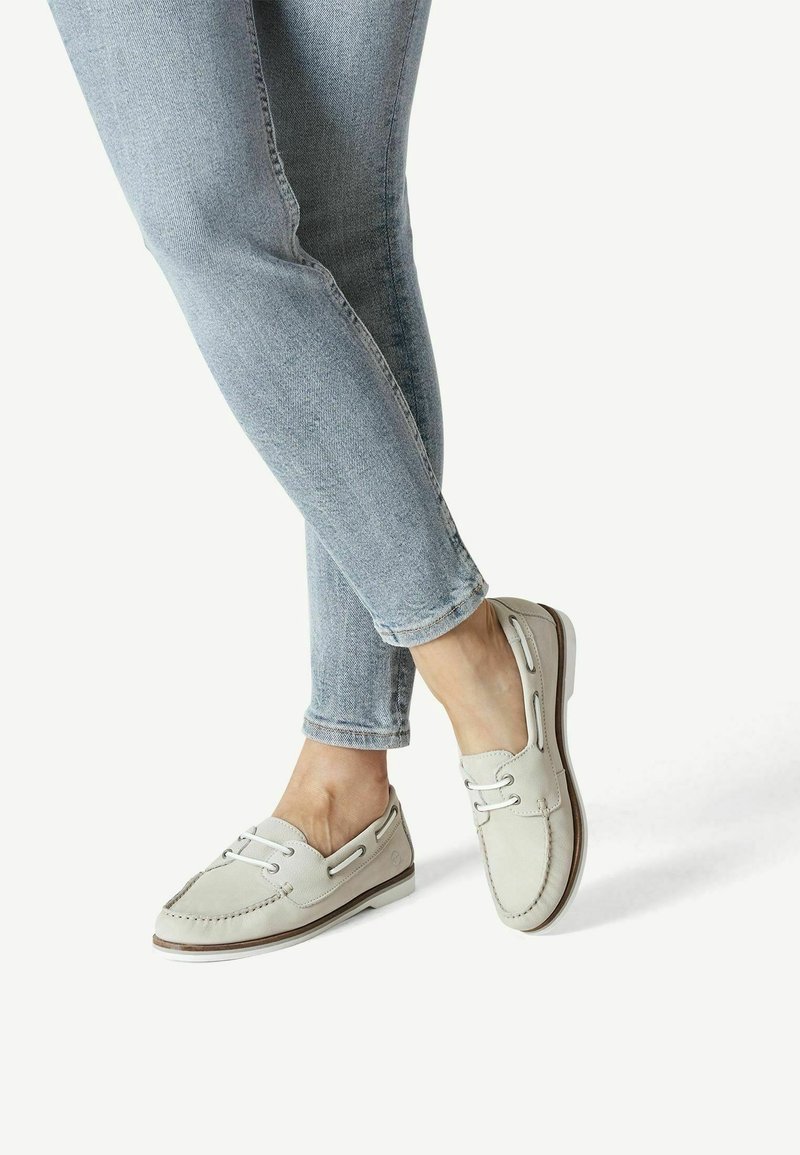 Bateaux légers gris clair avec une surface en cuir texturé, lacets blancs et semelle brune contrastante, associés à un jean bleu clair.