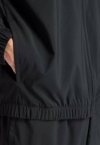 Schwarze Windjacke mit elastischen Bündchen, durchgehendem Reißverschluss, Seitentaschen und gerafftem Saum, aus einem glatten, strukturierten Stoff.