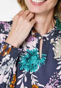 Bluse mit Blumenmuster, die aus dunkelblauem Stoff mit türkisfarbenen, lila und orangefarbenen Blumen besteht. Schlüssel-Ausschnitt-Detail und geraffte Ärmelabschlüsse hervorgehoben.